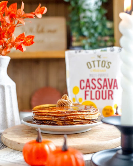 Pumpkin-spice thin pancakes in a stack on a plate. Topped with cinnamon butter. Bag of Otto's Naturals Cassava Flour in background, and little plastic pumpkins in foreground