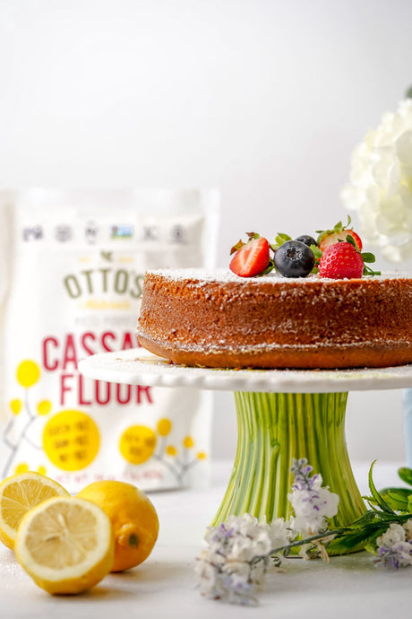 Grain-Free Olive Oil cake on cake stand, with fresh berries and powdered sugar on top. Bag of Otto's Naturals Cassava Flour in background and lemons on counter