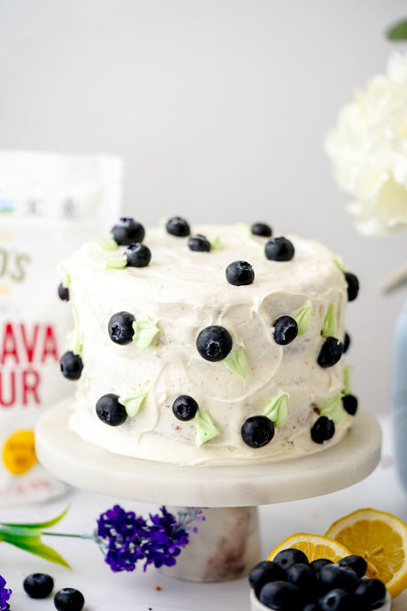 Blueberry layer cake on cake stand with flowers and lemons on counter. Otto's Naturals Cassava Flour bag in background