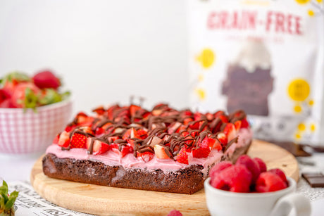 Grain-Free tray of brownies with strawberry buttercream, fresh chopped strawberries, and drizzled chocolate on top. Bag of Otto's Grain-Free Classic Brownie mix in background