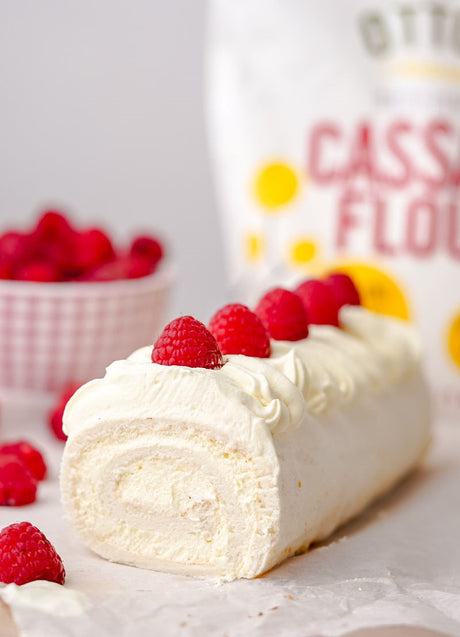 White grain-free swiss roll filled with whipped cream, and topped with whipped cream and fresh raspberries. Bowl of raspberries and bag of Otto's Naturals Cassava Flour in background 