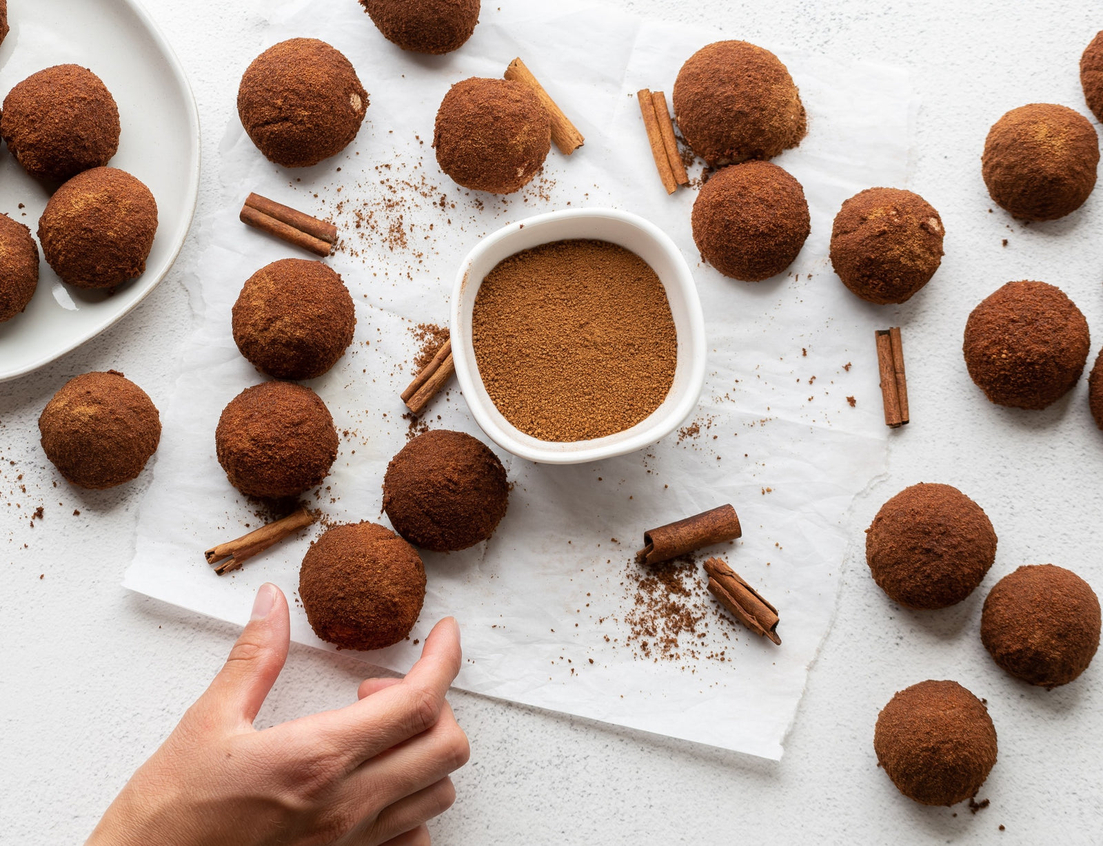 Hand reaching for grain-free cinnamon-sugar donut hole. Donut holes lying on counter with bowl of cinnamon-coconut sugar in the center, and some broken cinnamon sticks nearby