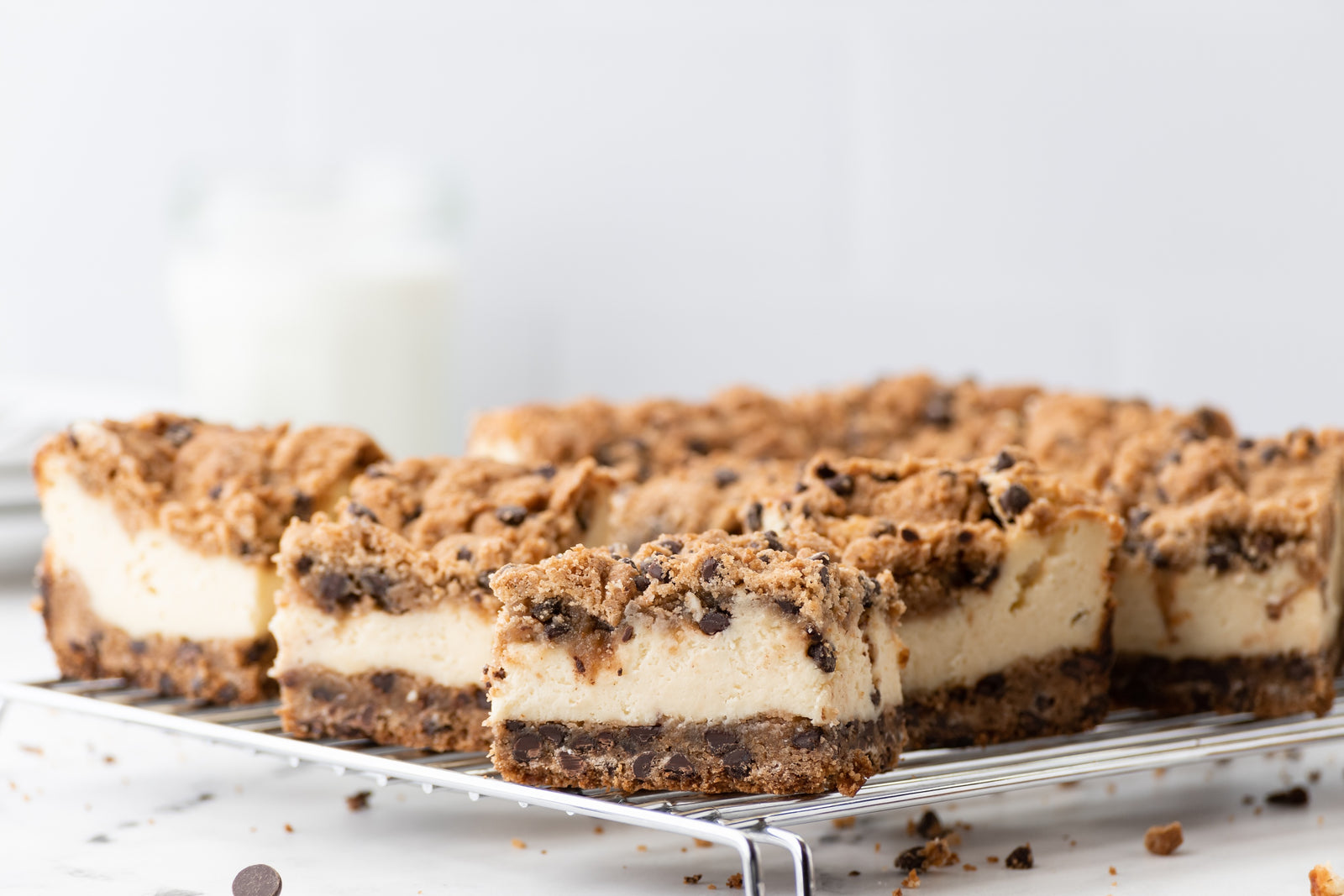 wire rack with squares of grain-free cookie-cheesecake bars. Base and topping is chocolate chip cookie, while the middle layer is cheesecake.