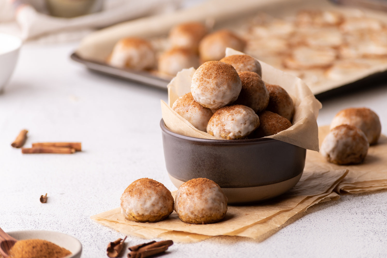 bowl of baked grain-free pumpkin donut holes, with tray of donut holes and glaze in background. More doughnut holes and fall spices sitting on counter