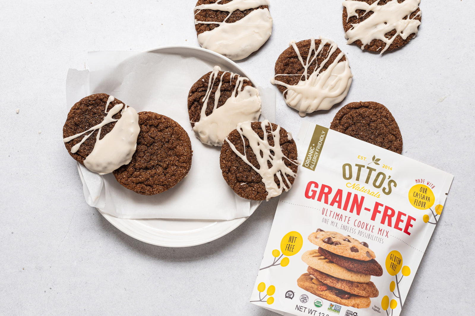 grain-free gingerbread cookies with icing on plate and on counter. Bag of Otto's Naturals Grain-Free Ultimate Cookie Mix lying flat on counter