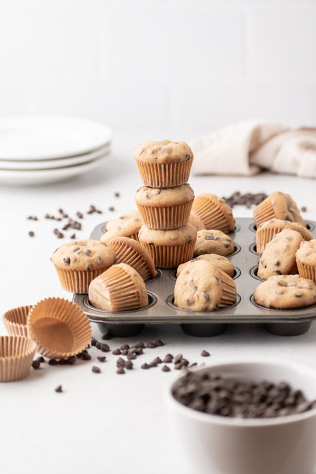 mini grain-free chocolate chip muffins in a mini muffin tin, with 3 muffins stacked above the others. bowl of chocolate chips, cupcake wrappers, and plates on counter nearby