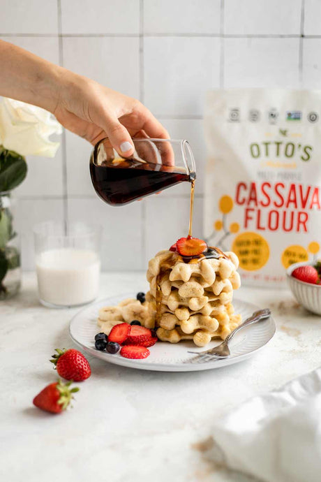 Fluffy Grain-Free Waffles stacked on a plate with berries. maple syrup being poured on top and Otto's Cassava Flour in background